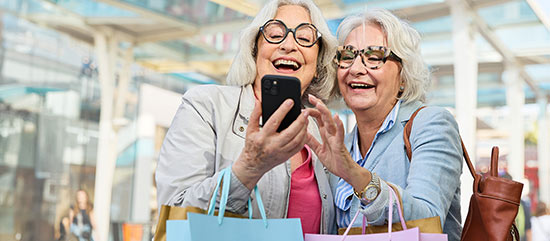 Zwei Frauen nutzen beim Einkaufen gemeinsam digitale Angebote auf einem Smartphone.