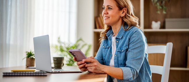 Happy mature woman working from home using a laptop and smartphone, smiling while looking away from the screen